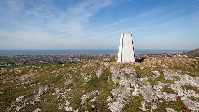 Trig point on Graig Fawr beside rocky terrain, with a view of towns and the sea in the background.